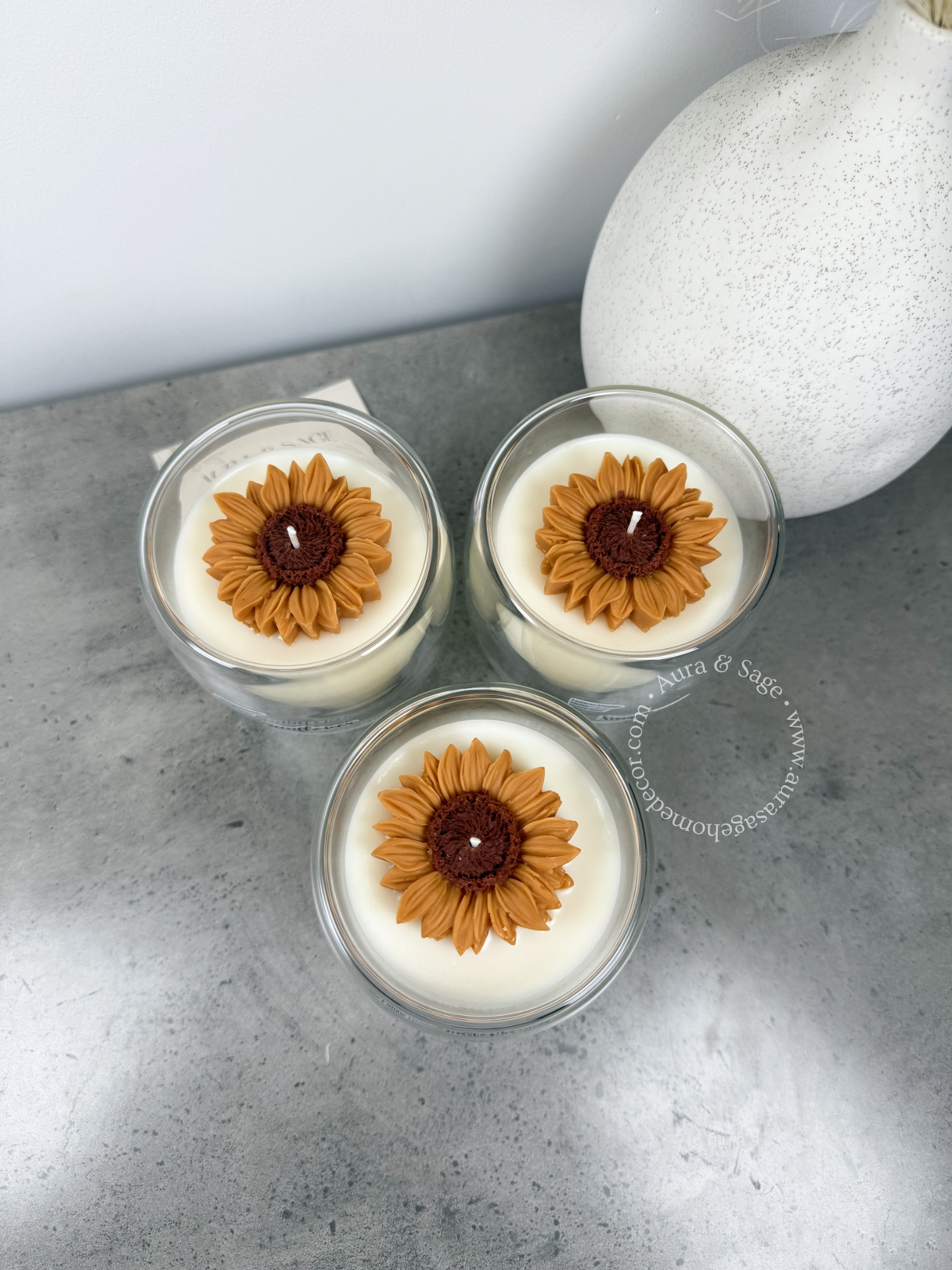 Three sunflower-themed candles in glass containers on a gray surface.