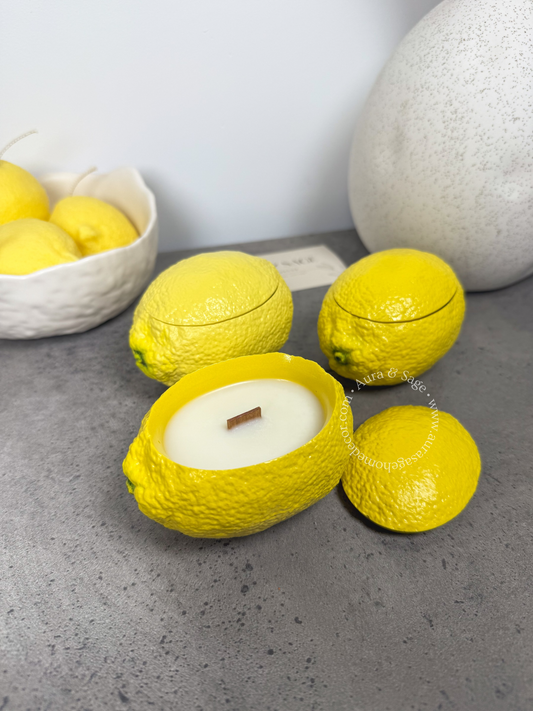 Yellow lemon-shaped candles on a gray surface with a bowl of lemons in the background.